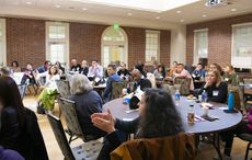 A large group of people seated in a room arranged with tables, some engaged in conversation while others appear to be listening attentively.
