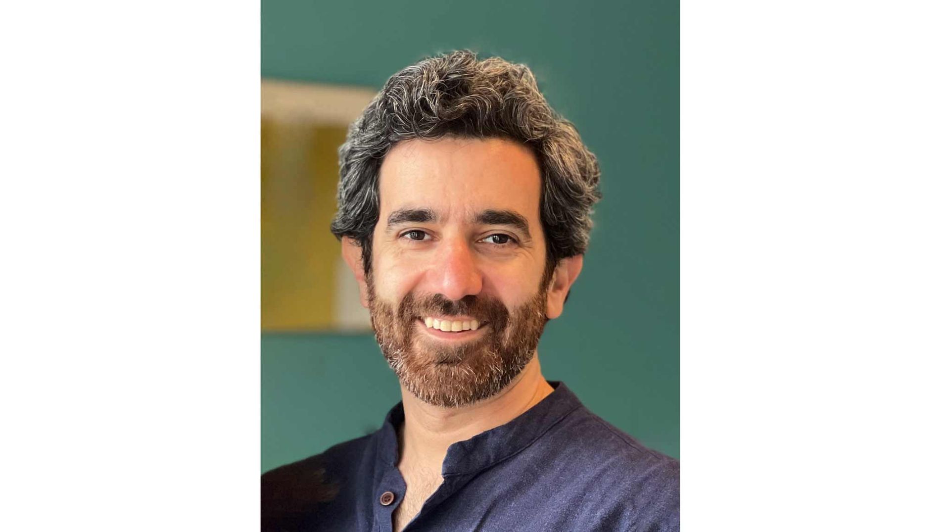 The head and shoulders of a man with a beard standing in front of an out of focus green wall.