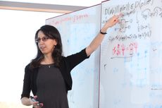 Nicole Yunger Halpern stands looks to the left while pointing to one of several physics equations on the blackboard behind her.