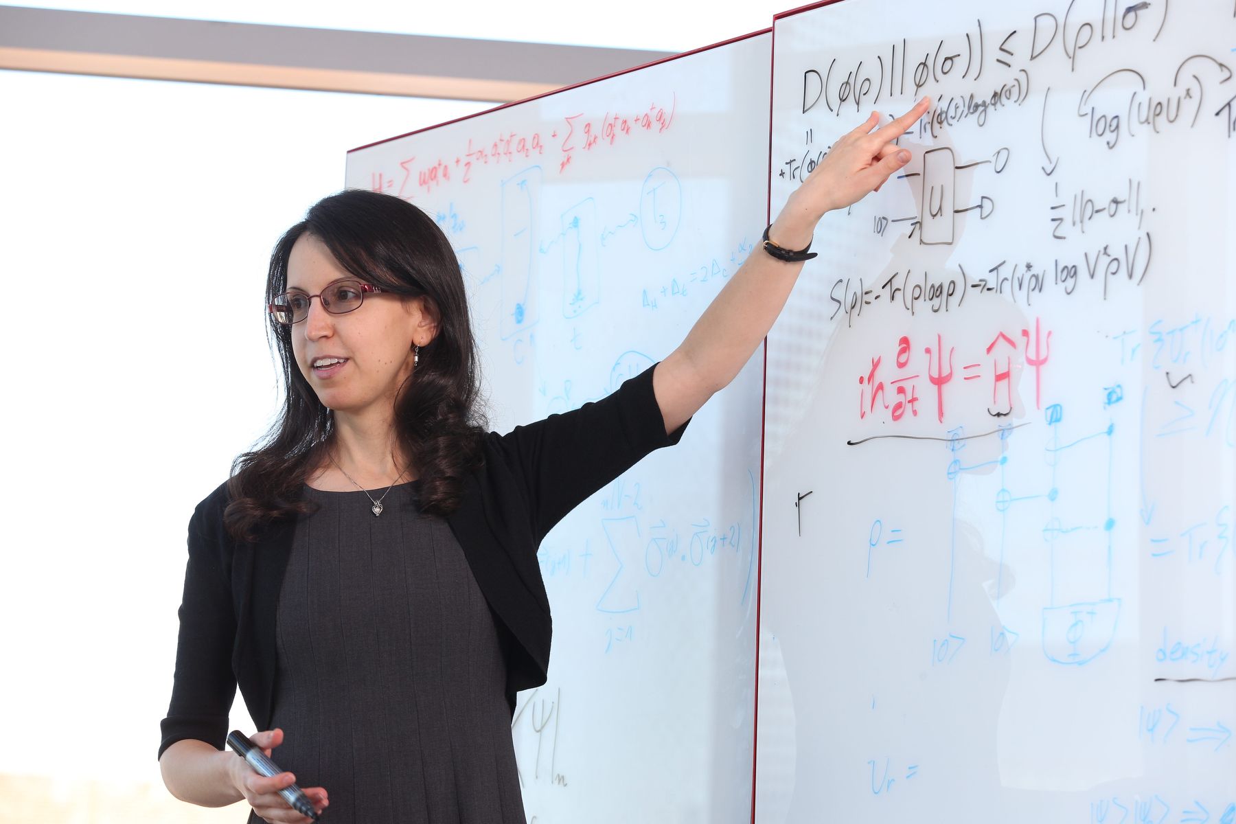 Nicole Yunger Halpern stands looks to the left while pointing to one of several physics equations on the blackboard behind her.